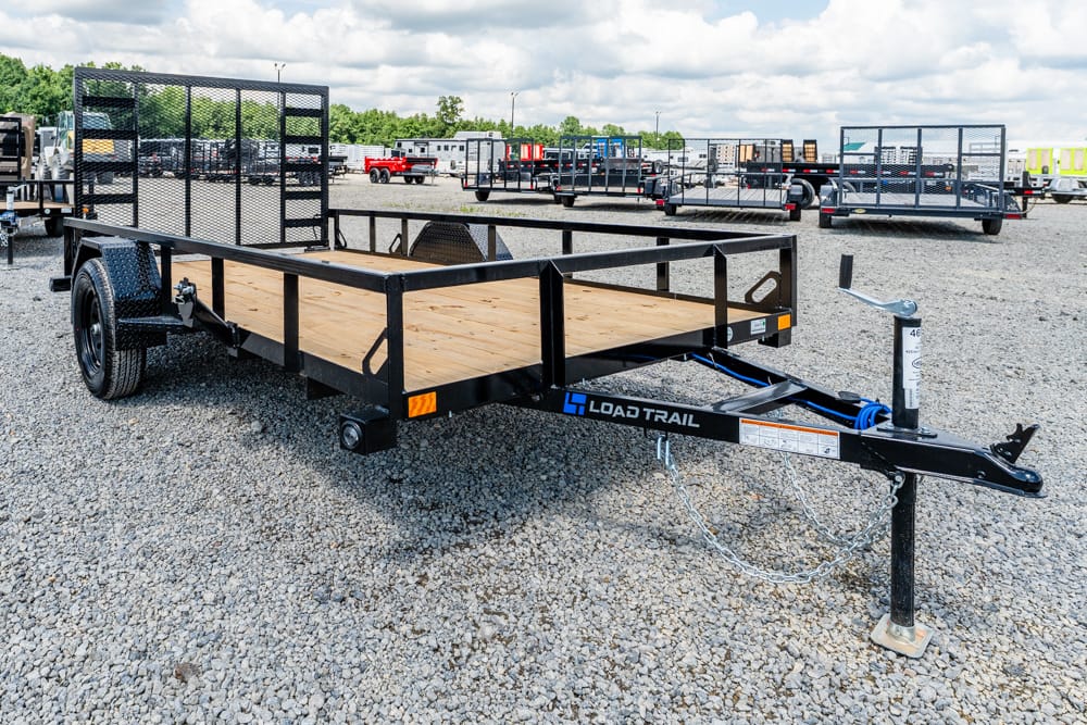Two-axle Load Trail utility trailer with a wooden deck and side rails, parked on a gravel lot among other trailers.