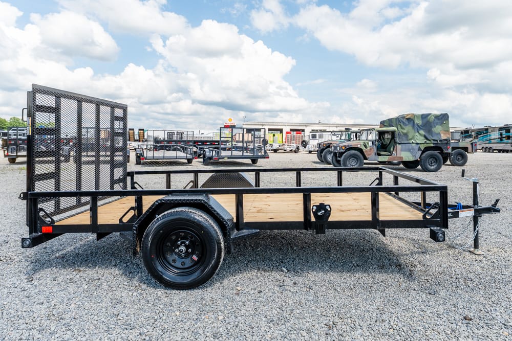 Black flatbed utility trailer with a wooden deck sits on a gravel lot, with other trailers and a military vehicle in the background.