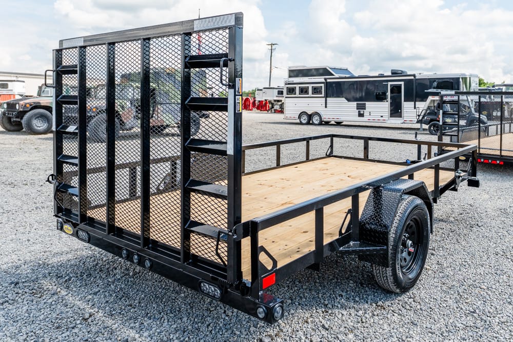 Black utility trailer with wood deck and tall mesh ramp gate in a gravel lot, ready to haul equipment