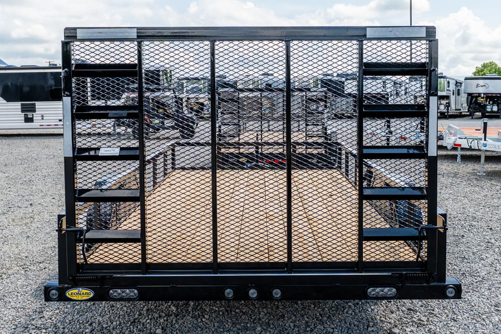 Black utility trailer with a metal mesh ramp gate and wooden bed, parked on a gravel lot with other trailers in the background (Leonard logo on front).