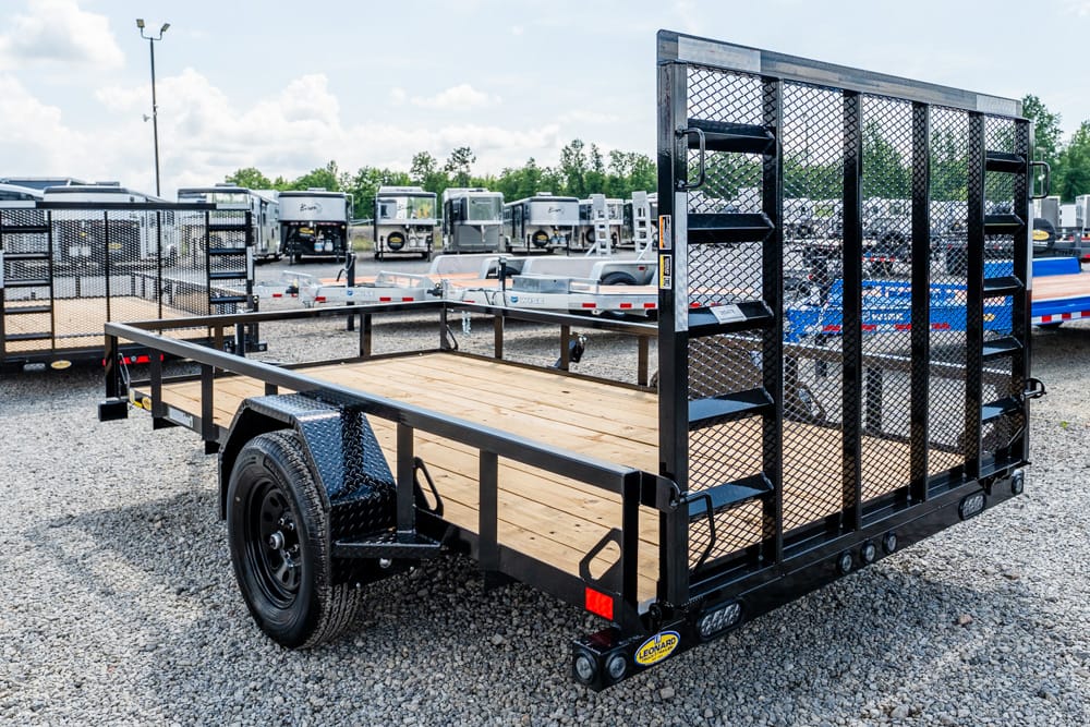 Metal flatbed utility trailer with a wooden deck and tall mesh rear ramp in a gravel lot, ready for hauling equipment or vehicles.