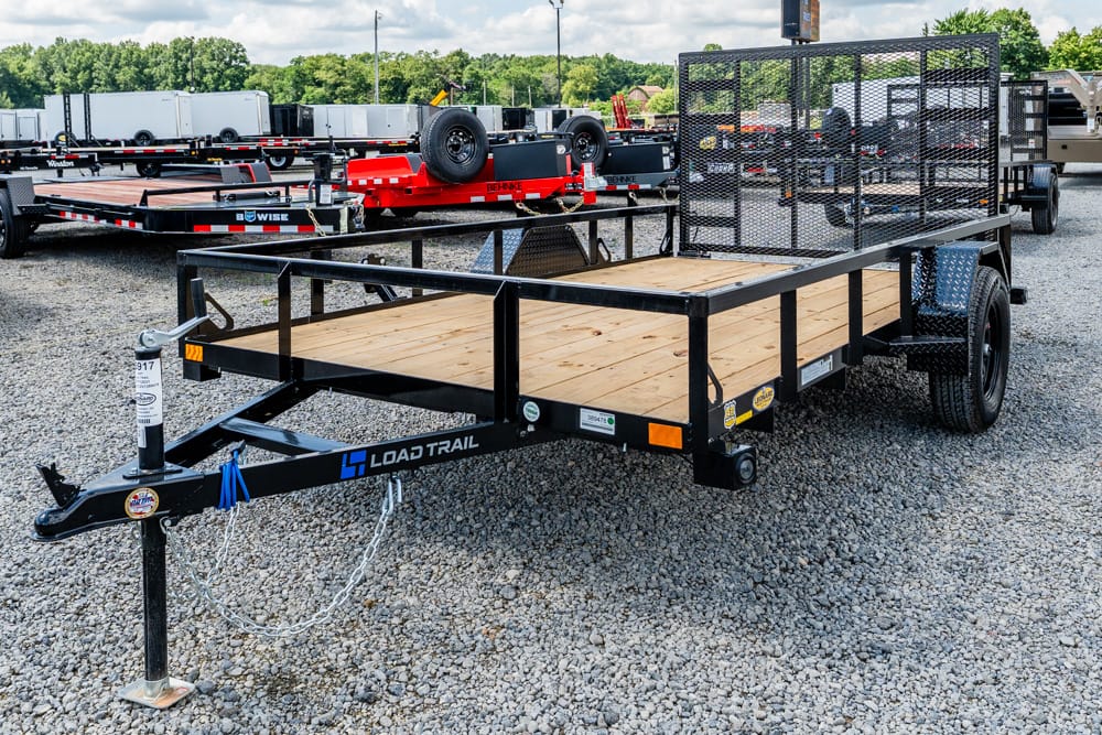 Load Trail flatbed trailer with a wooden deck, dual axles, and a mesh rear gate on a gravel lot.