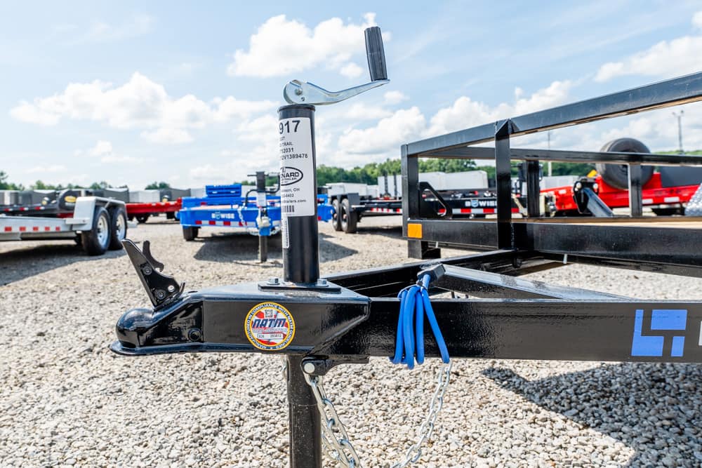 Close-up of a black trailer hitch on a gravel lot, with a blue safety chain and unit jack in the foreground and other trailers in the background.