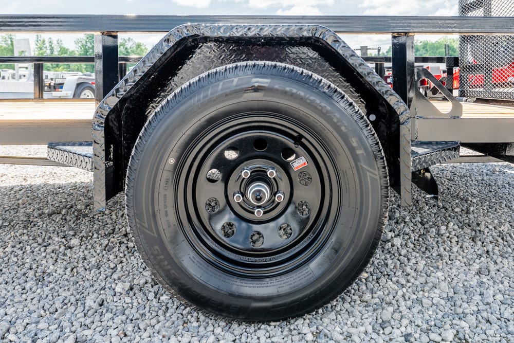 Close-up of a black steel trailer wheel and tire mounted on a diamond-plate fender, with gravel ground behind.