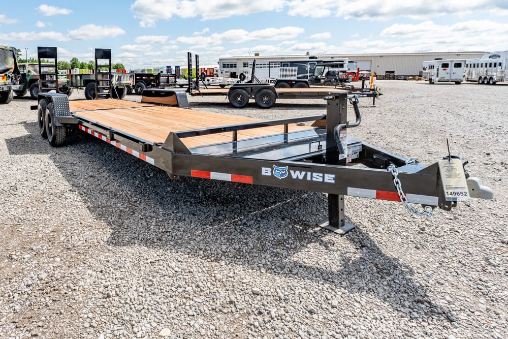 Black B-WISE flatbed trailer with a wooden deck parked on a gravel lot, with other trailers and a warehouse in the background.