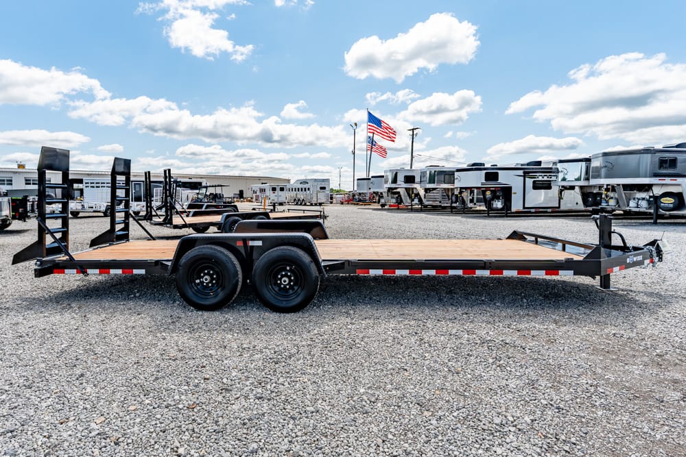 Flatbed trailer with dual axles and wooden deck parked on a gravel lot, with RVs and American flags in the background.