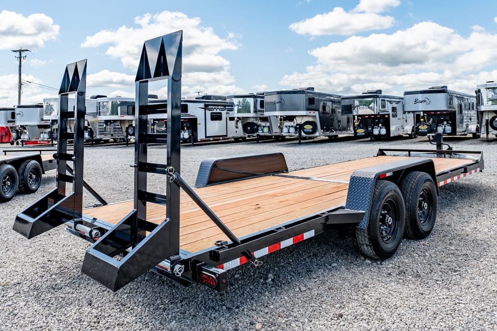 Flatbed trailer with a wooden deck and folding ramps in a gravel RV lot on a sunny day.
