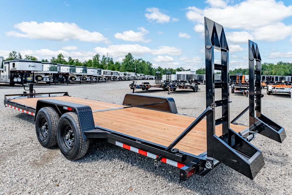 Flatbed car trailer with dual loading ramps raised at the back, wooden deck, two axles, parked on a gravel lot in a trailer yard.