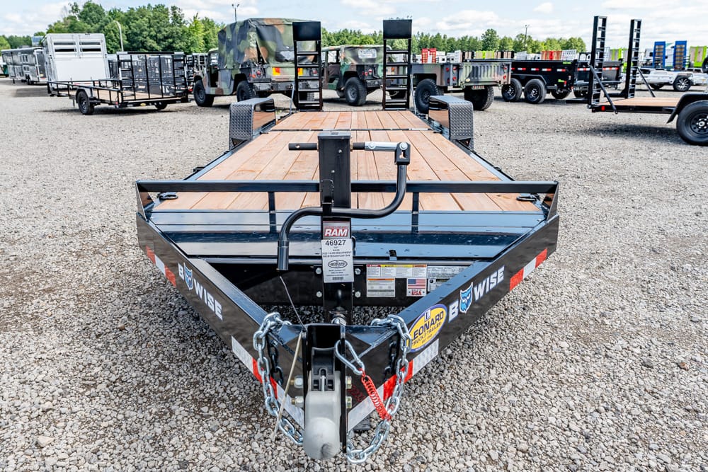 Front view of a black steel utility trailer with a wooden deck in a gravel lot, showing hitch, chains, and RAM label.