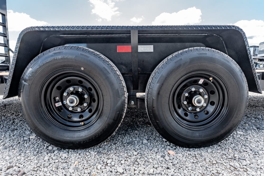 Dual-axle trailer wheels with black rims on a gravel surface under a blue sky.