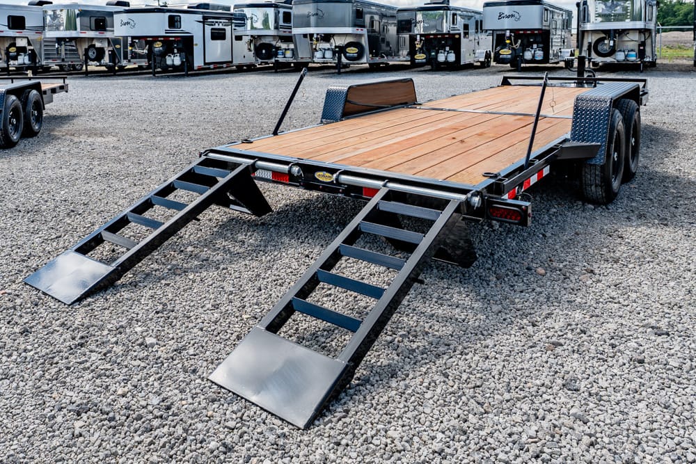 Flatbed trailer with two fold-down loading ramps on a gravel lot, ready for vehicle loading