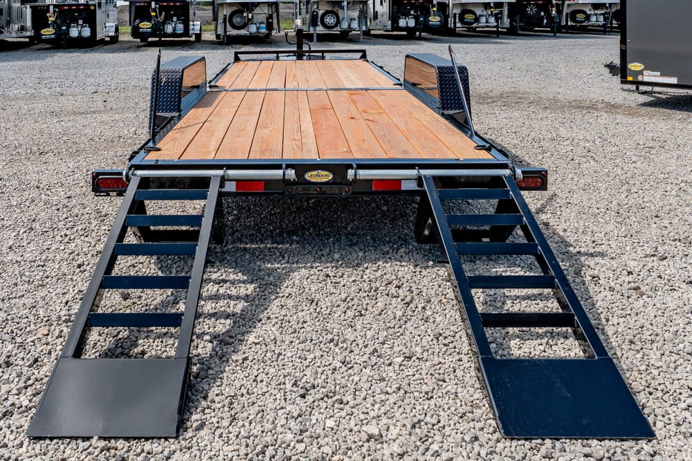 Flatbed trailer with a wooden deck and two metal loading ramps extended on a gravel lot.