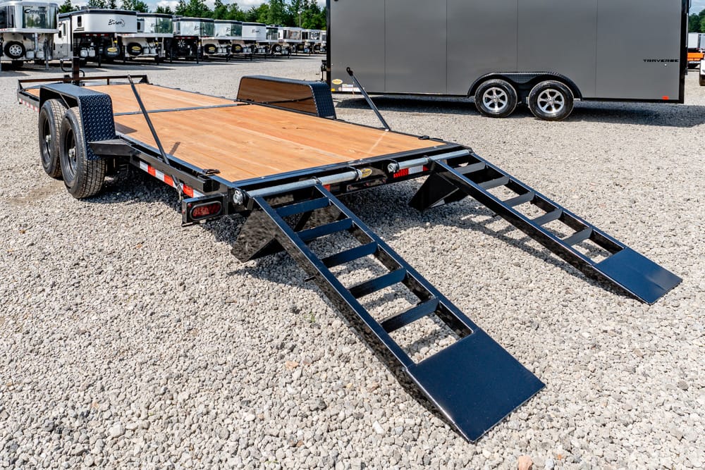 Flatbed trailer with wooden deck and two folding loading ramps on a gravel lot used for vehicle transport/haulage.