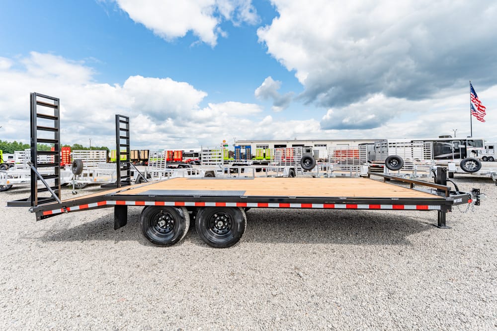 Flatbed trailer with dual rear wheels and wooden deck parked in a gravel lot with other trailers nearby.
