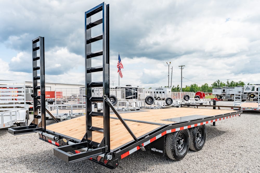 Black steel flatbed trailer with a wood deck and dual tall loading ramps, parked in a gravel lot with other trailers and an American flag in the cloudy sky background.