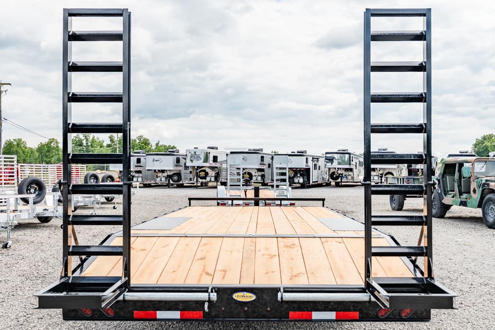 Wide shot of a black steel flatbed trailer with tall upright ramps, wooden deck, in a gravel lot with RVs and vehicles in the background.