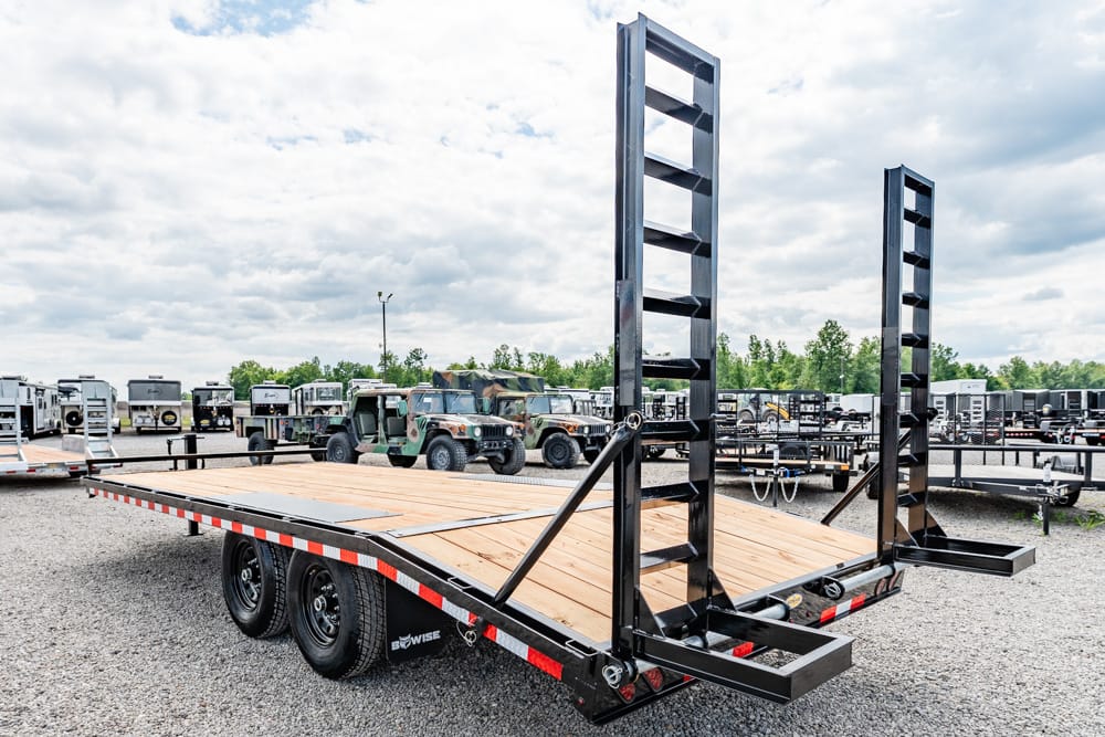 Flatbed trailer with dual fold-up ramps in a gravel lot, surrounded by other trailers and vehicles.