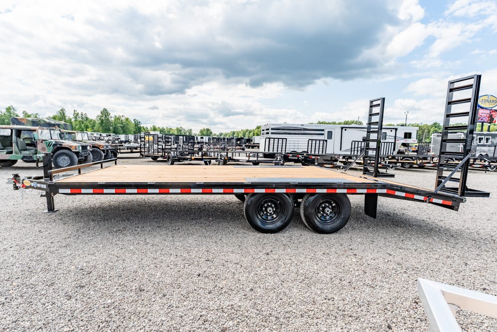 Flatbed trailer with a wooden deck and ramps, parked on a gravel lot among other trailers and trucks under a cloudy sky.