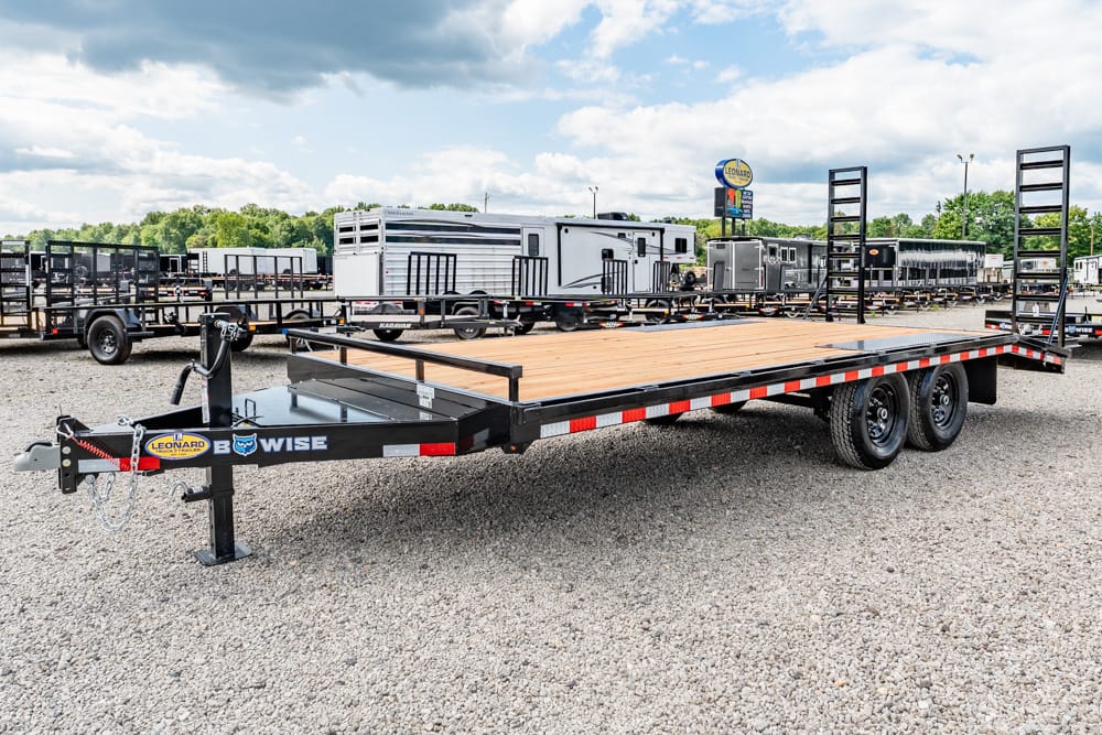 Black flatbed trailer with a wooden deck and dual axles, parked on a gravel lot with other trailers and RVs in the background, ramps raised at the rear.