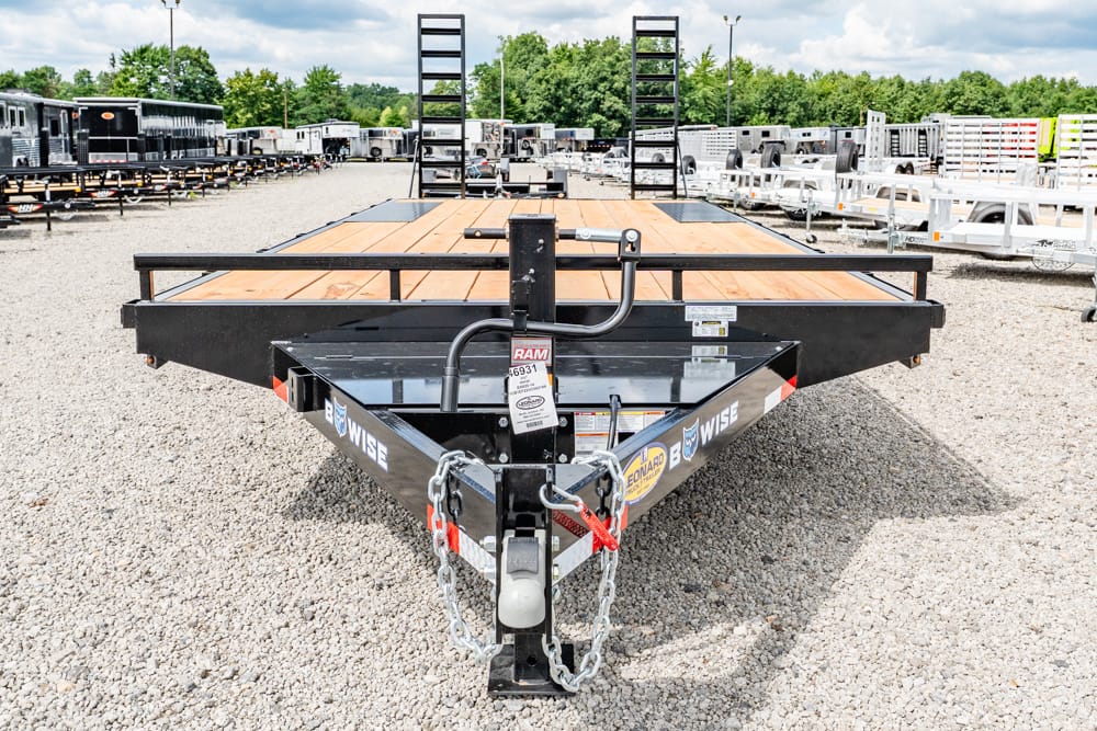 Front view of a black flatbed trailer with a wooden deck, parked on a gravel lot among many trailers, with safety chains and hitch visible.