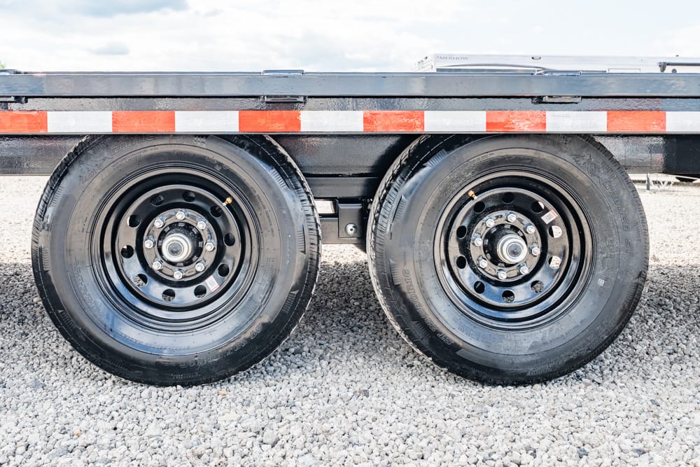 Close-up of two black trailer wheels with metal rims on gravel beneath a flatbed trailer with red-and-white reflective stripes.