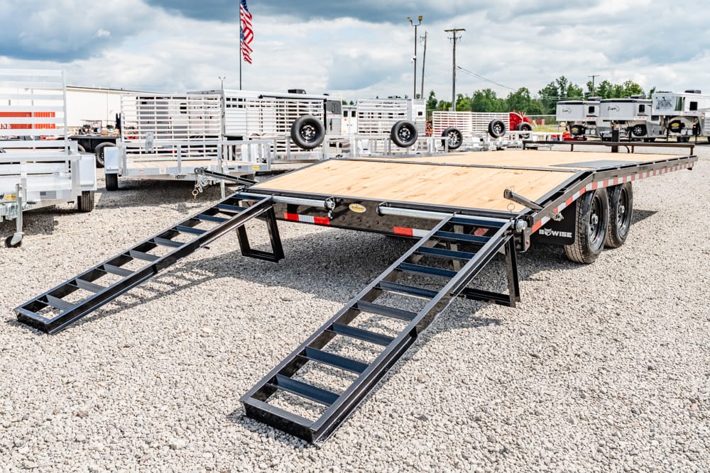 Flatbed trailer with two metal loading ramps extended on a gravel lot; other trailers in the background on display.