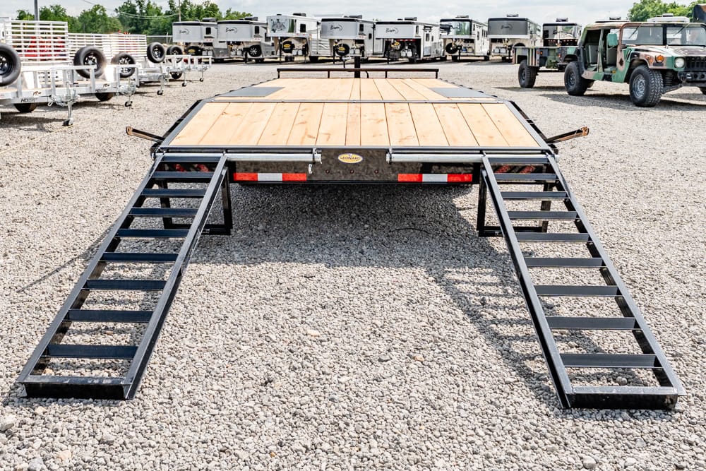 Flatbed trailer with two metal ramps extended on a gravel lot, surrounded by trailers and trucks in the background.