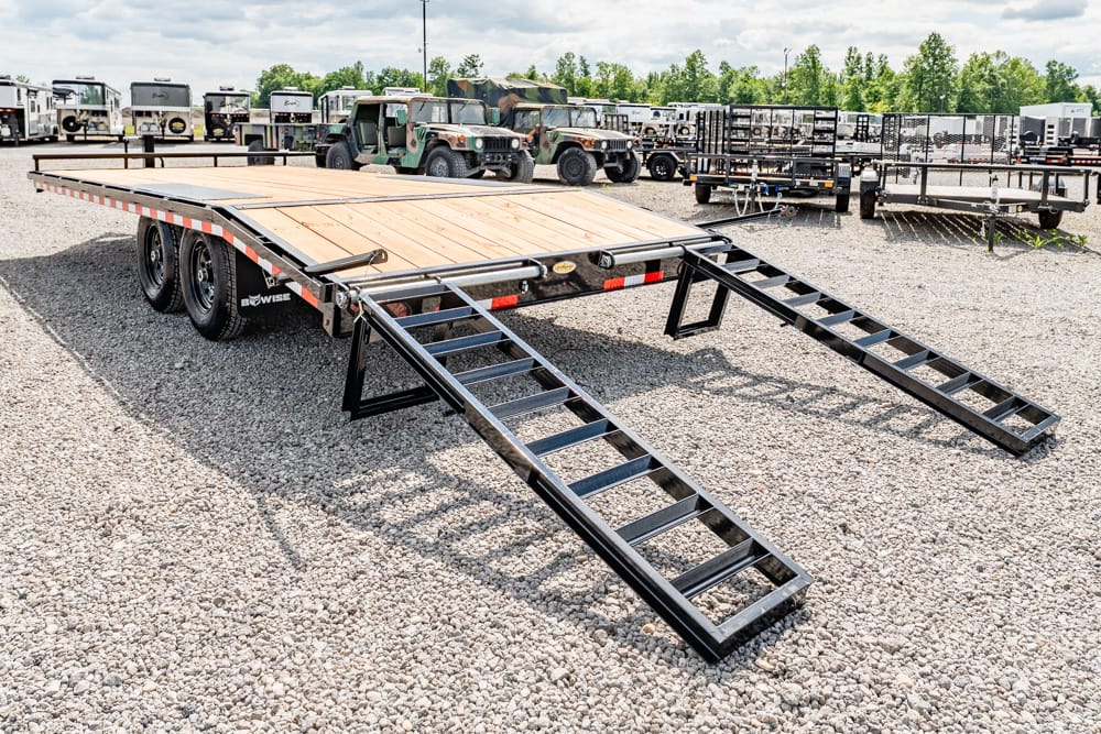 Flatbed trailer with a wooden deck and two metal ramps extended on a gravel lot, surrounded by other trailers.