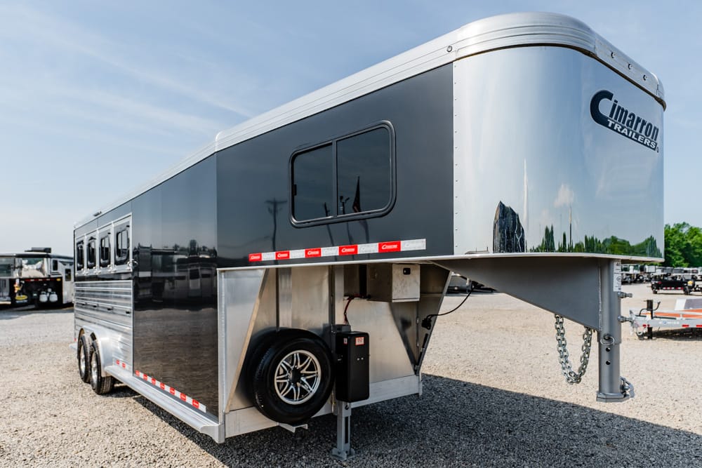 Gooseneck horse trailer with black-and-silver exterior, spare tire mounted under front, Cimarron Trailers logo, parked on gravel.