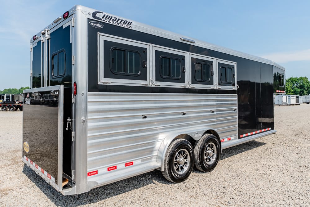 Silver and black horse trailer with multiple windows and two axles, parked on gravellot. The trailer has a rear awning door open and chrome wheels.