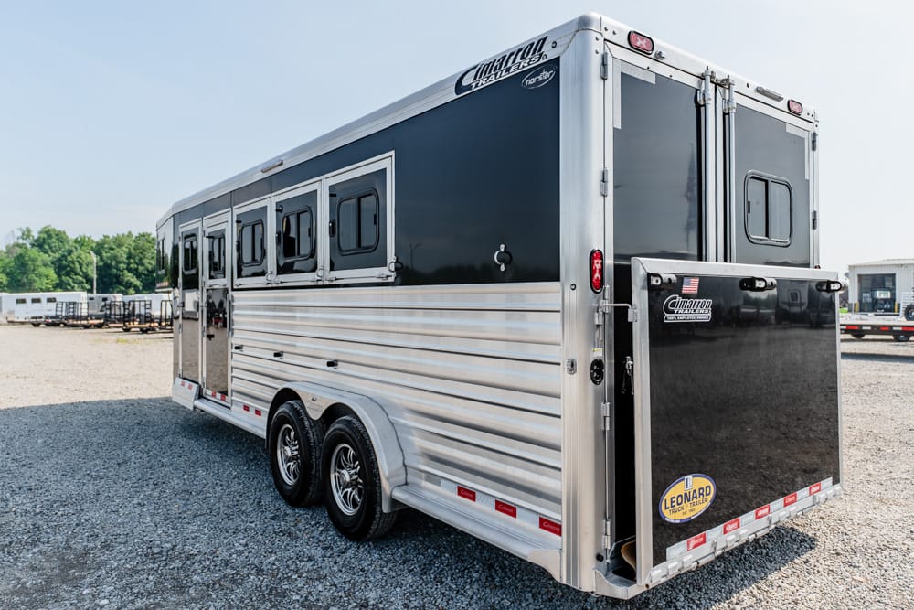 Silver and black double-axle horse trailer with multiple side windows on a gravel lot, rear ramp closed, Cimarron Trailers branding visible.