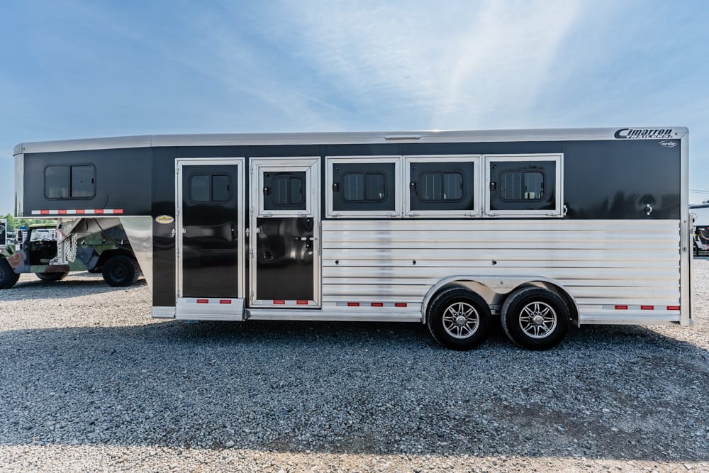 Silver-and-black horse trailer with four side windows and dual axles, parked on gravel under a blue sky (Cimarron Trailers logo visible).