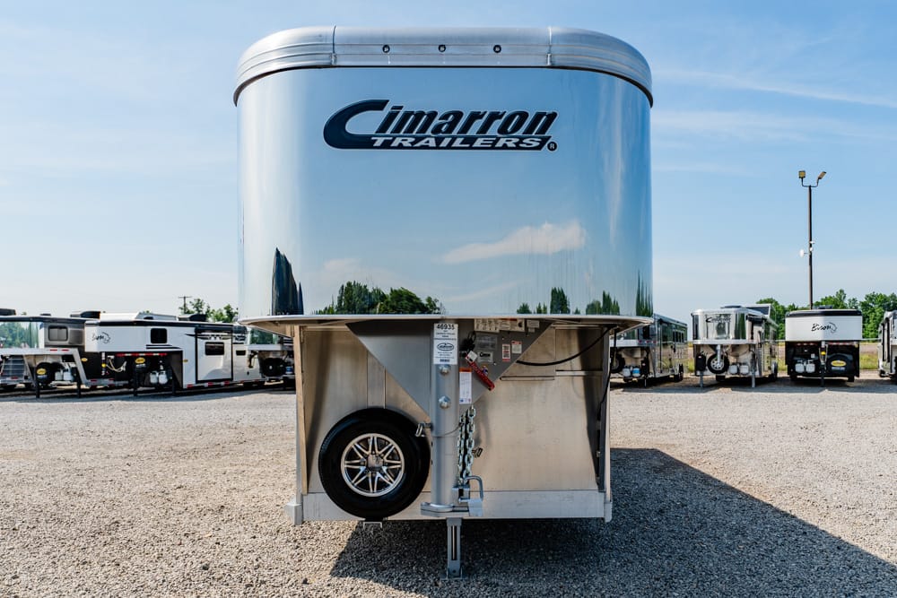 Polished aluminum Cimarron gooseneck trailer with a spare tire at the front, parked in a lot with other trailers.