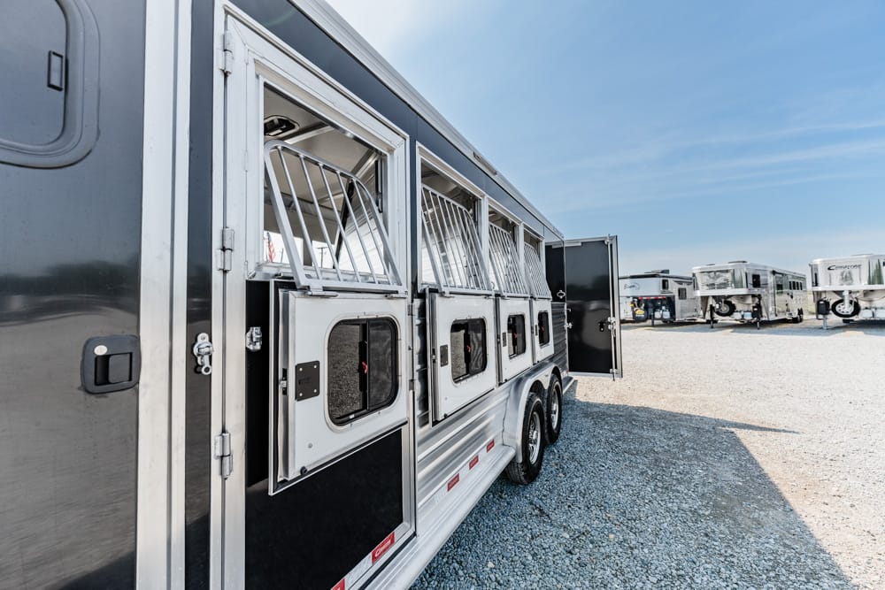 Silver horse trailer with open side compartments showing stalls, parked on a gravel lot with other trailers in the background on a sunny day.