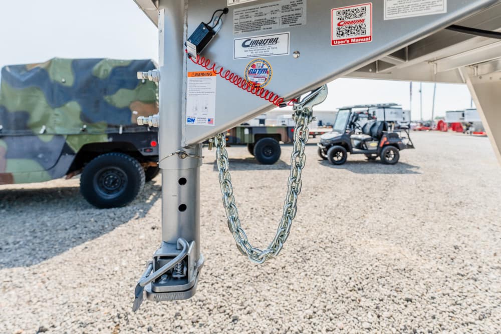Close-up of a trailer hitch with safety chains hanging from the hitch, in a gravel yard with utility vehicles in the background.
