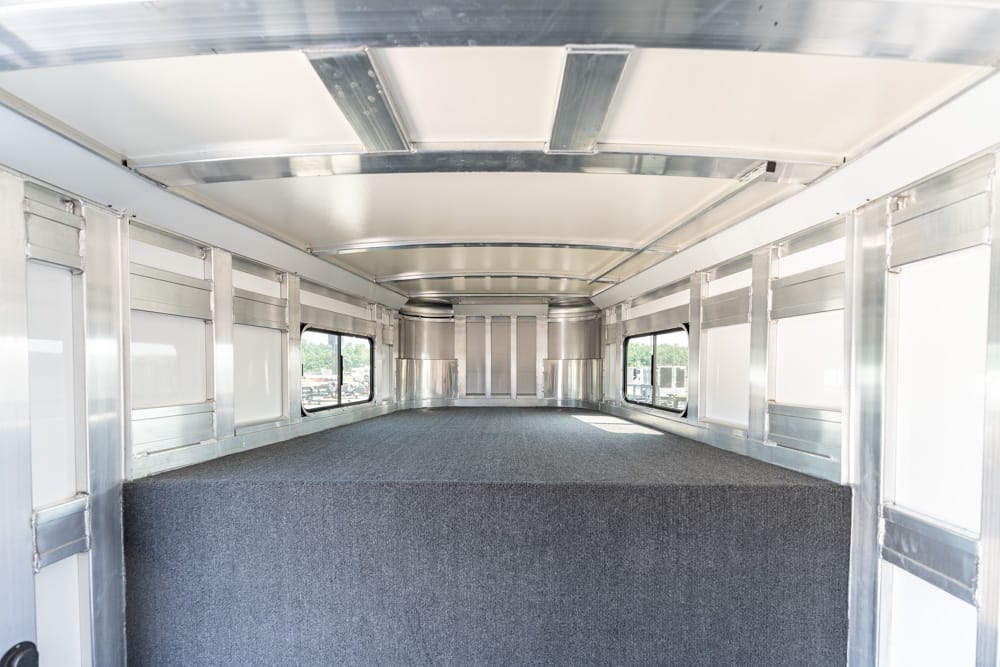 Interior of a metal trailer with a raised gray carpeted platform and side windows. Empty cargo space inside a shiny aluminum trailer.