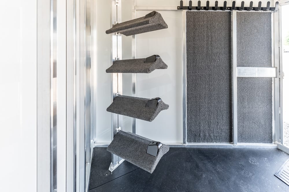 Inside a horse trailer: four foam-padded saddle racks on the left wall, with a textured rubber floor and a ramp/door on the right.