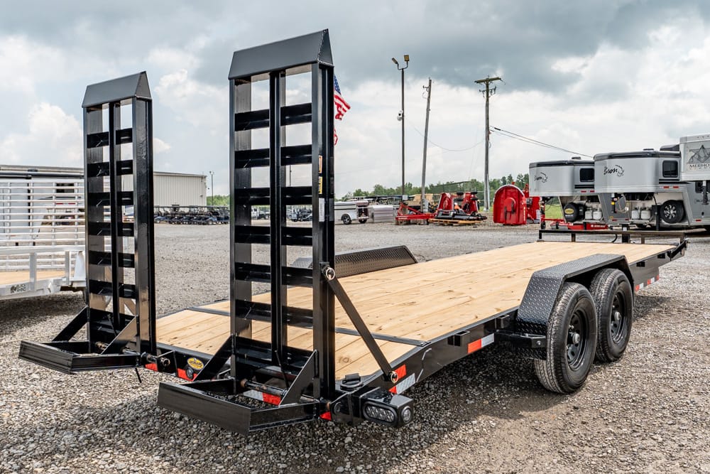 Black equipment trailer with wooden deck and tall dual ramps raised at the rear, parked on a gravel lot.