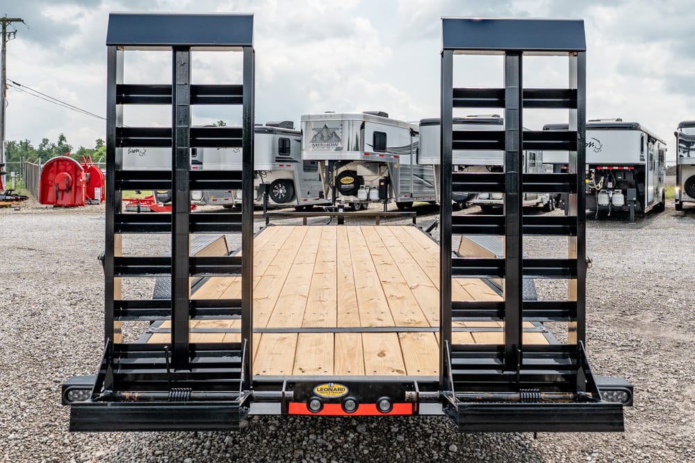 Back view of a black dual-ramp car hauler trailer with a wooden deck, in a gravel lot with RVs in the background.