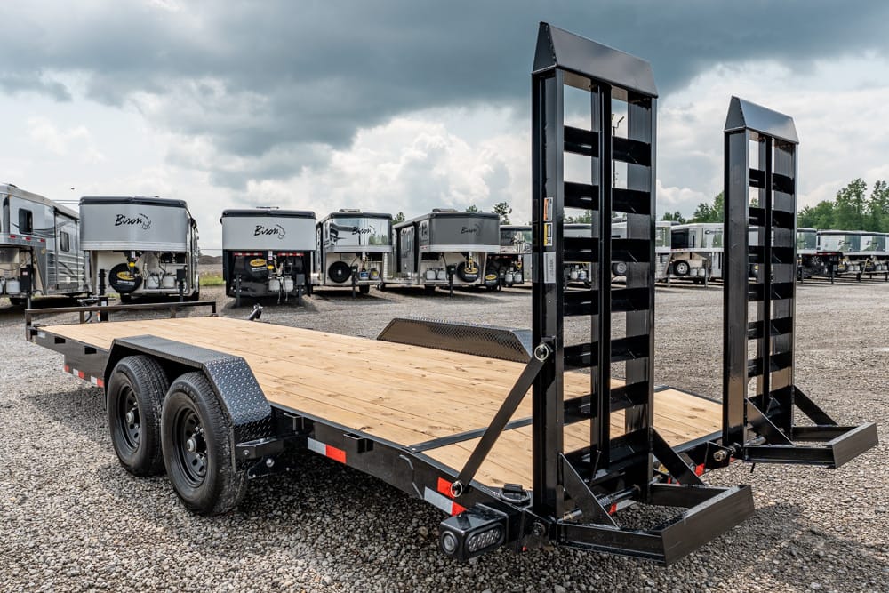 Flatbed trailer with wooden deck and tall fold-down ramps in a gravel lot, with several travel trailers in the background.