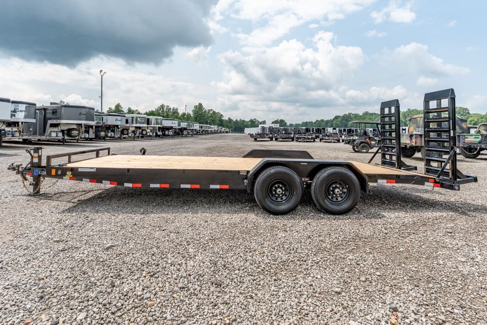Long flatbed trailer with dual axles and wood deck in a truck trailer yard, ramps up at the rear and space for hauling equipment or vehicles.