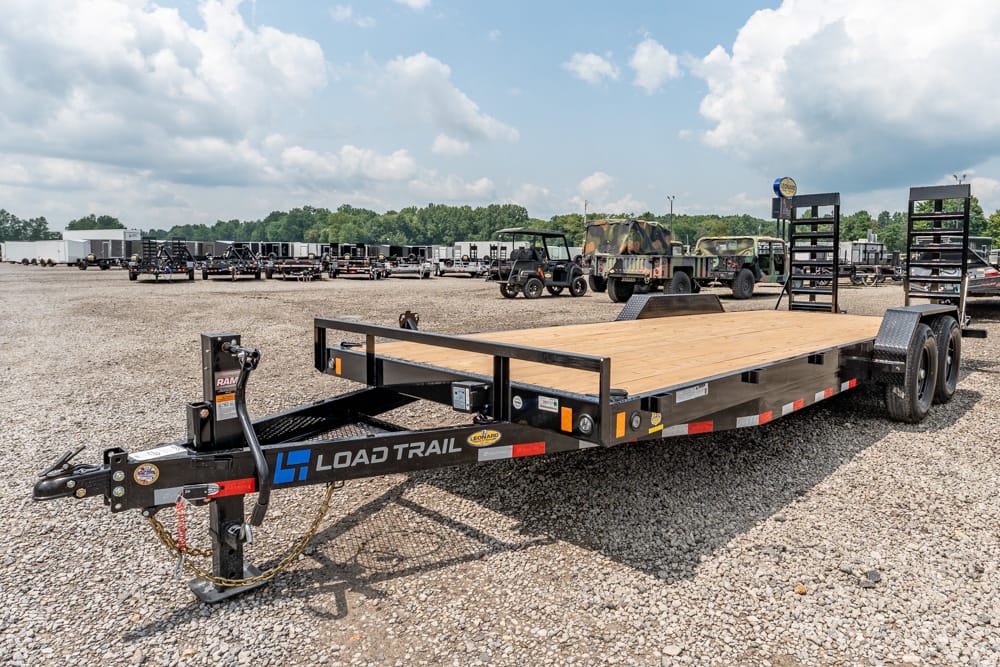 Load Trail flatbed trailer with wooden deck and dual axles parked on a gravel lot among other trailers and equipment