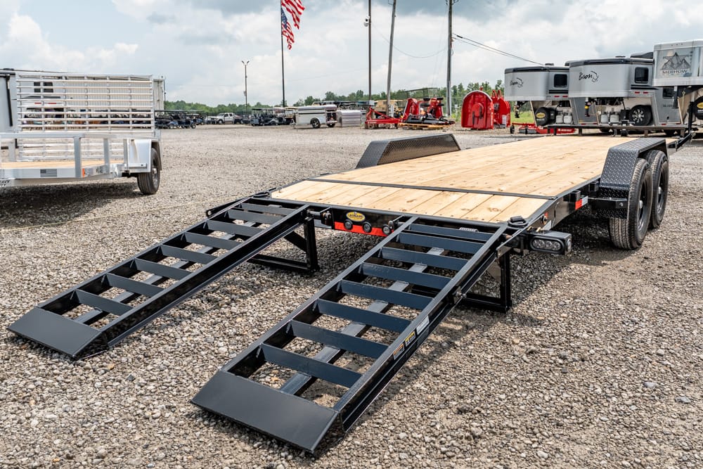 Flatbed trailer with a wooden deck and two black loading ramps extended, parked on a gravel lot with RVs in the background