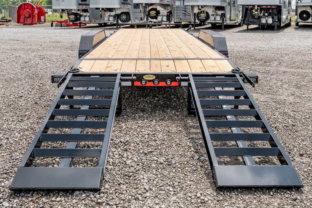Flatbed trailer with wooden deck and two metal loading ramps lowered onto a gravel lot.