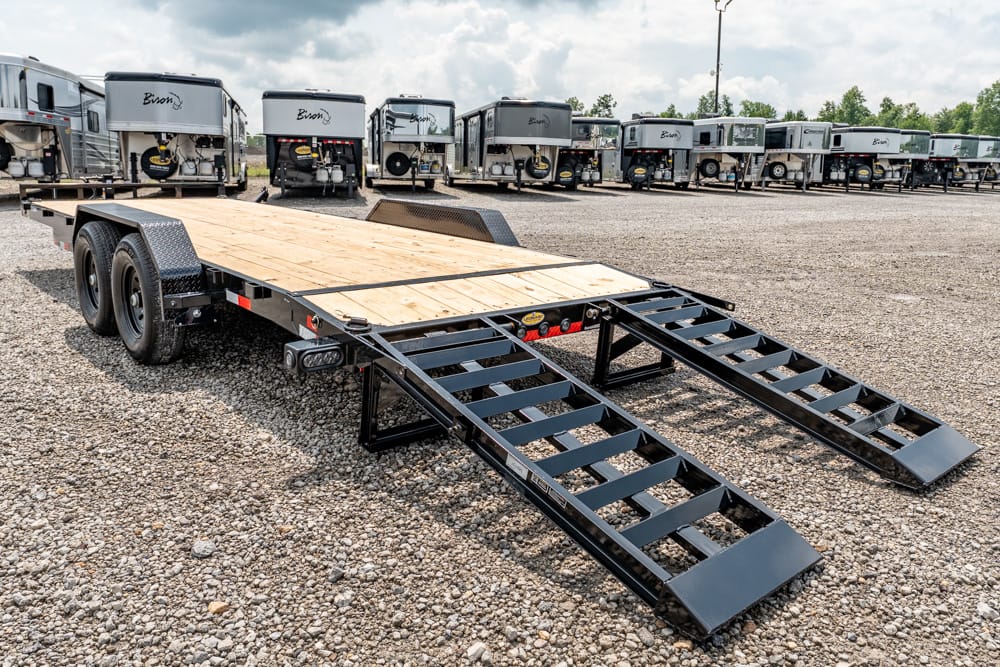 Flatbed trailer with two lowered loading ramps in a gravel lot, RV trailers lined up in the background