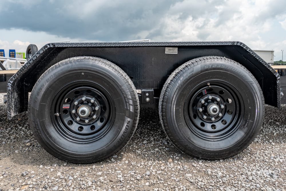 Dual-axle trailer with two black steel wheels on a gravel surface, under a metal fender.