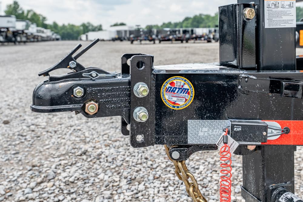 Close-up of a trailer hitch assembly with safety chains and latch on a gravel lot.