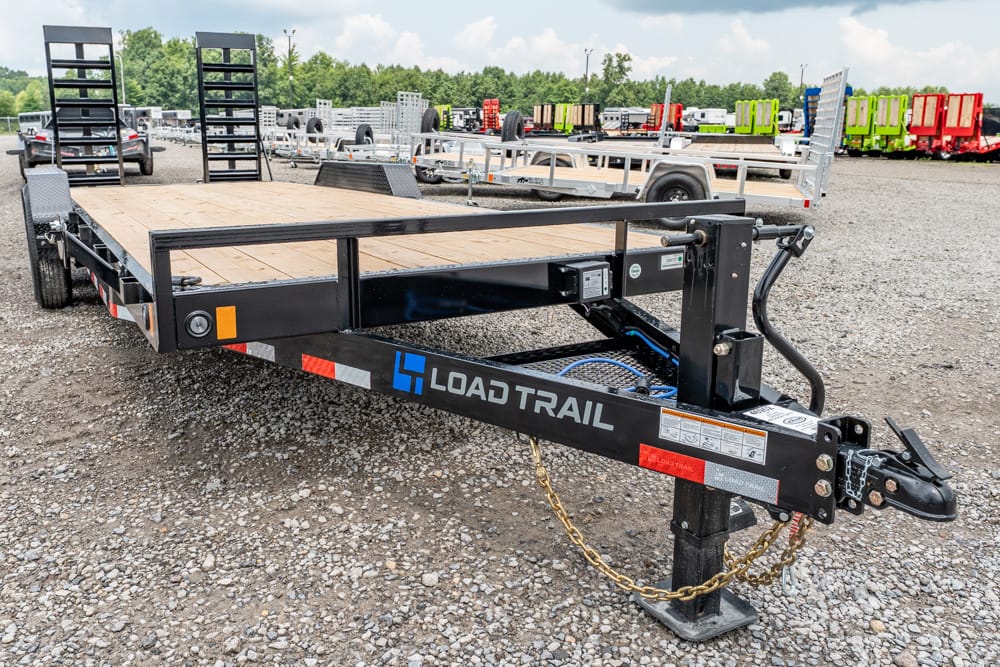 Black Load Trail utility trailer with a wooden deck on a gravel lot; visible hitch, safety chains, and rear ramps, with other trailers in the background.