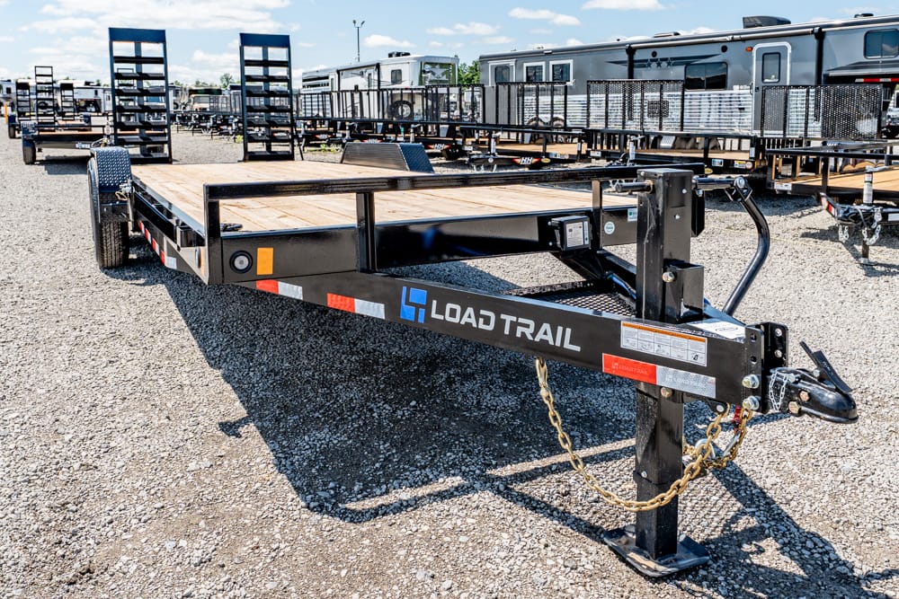 Front-right view of a Load Trail flatbed trailer with a wooden deck, parked on a gravel lot with other trailers in the background.