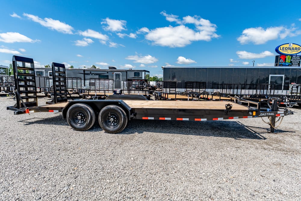 Flatbed trailer with dual axles parked on a gravel lot, with ramps at the rear and a bright blue sky above.